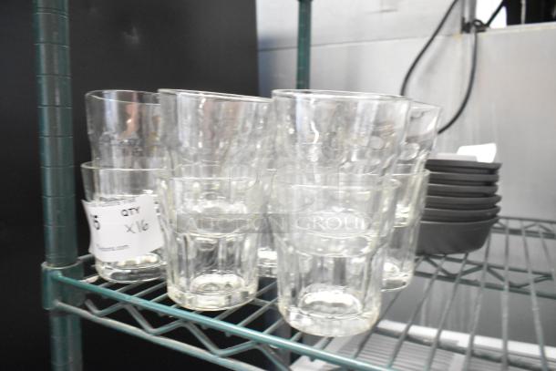 16 new beverage glasses on a shelf, some with minor scratches and dents. Includes a label reading "qty X16". 