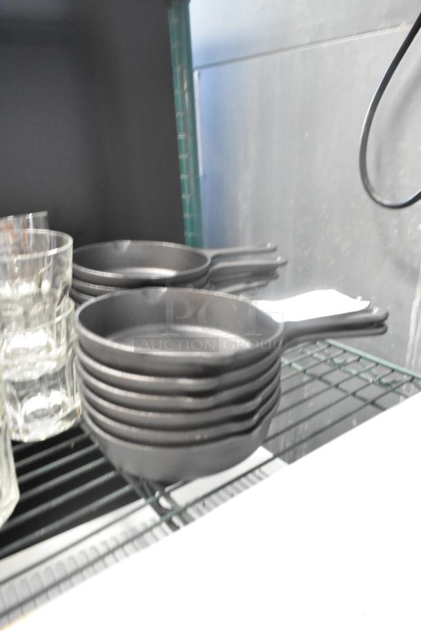 12 new Valor cast iron skillets, scratch and dent condition, stacked on shelves. Durable cookware, ideal for various heat sources.