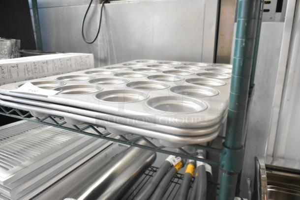 Three brand new metal 24-cup muffin baking pans, scratch and dent condition, stacked on a green metal shelf.
