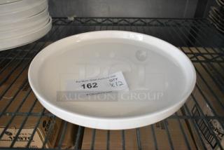12 new Acopa white plates, scratch and dent condition, on a metal shelf. Auction tag reads "162, QTY x12."