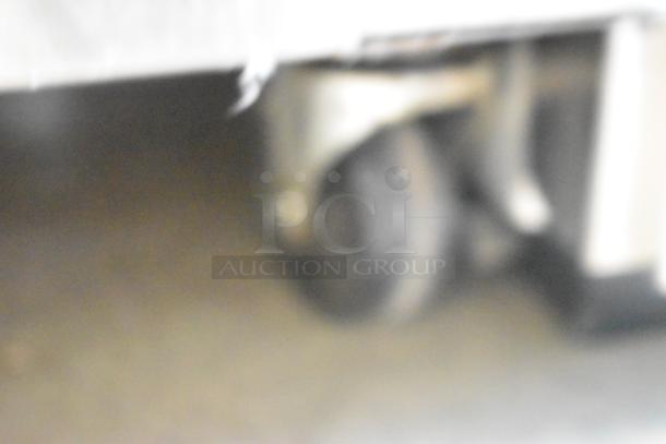 Blurry image of a metallic caster wheel, likely part of the Kool Star KSR49-S stainless steel cooler.