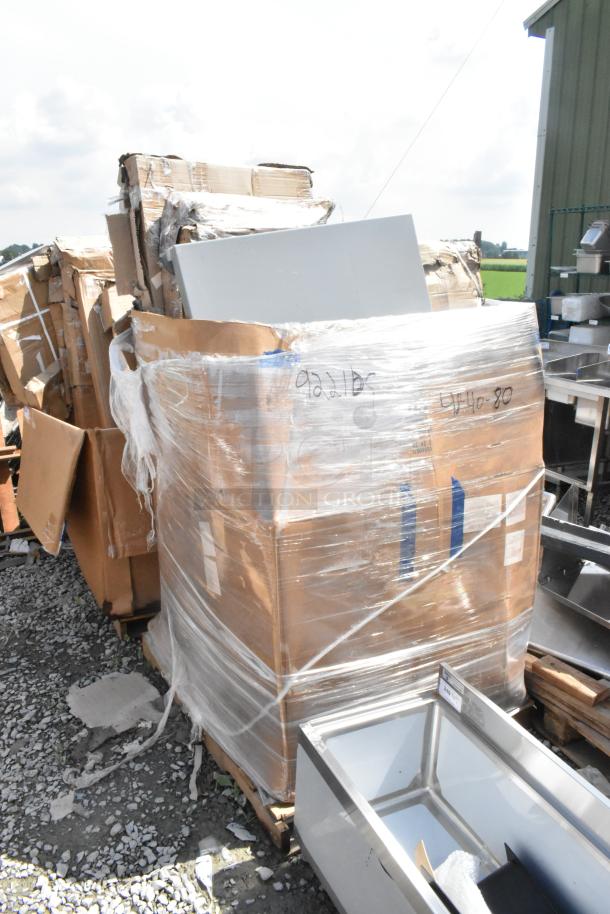 Plastic-wrapped pallets containing various new scratch and dent items, including a stainless steel table and platform truck, on a gravel surface.
