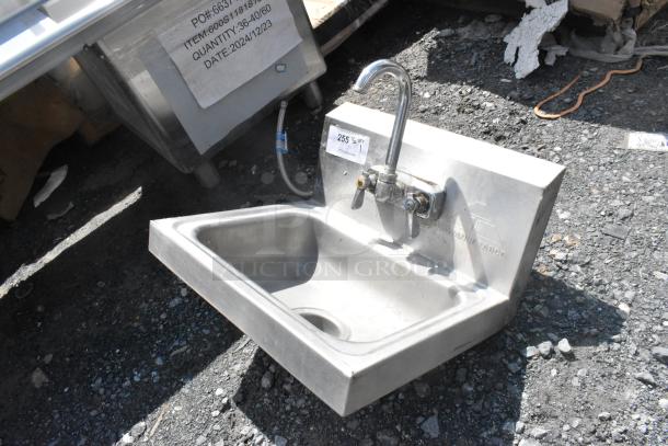 Stainless steel single bay wall mount sink with faucet and handles, shown outdoors, with visible part number label attached.