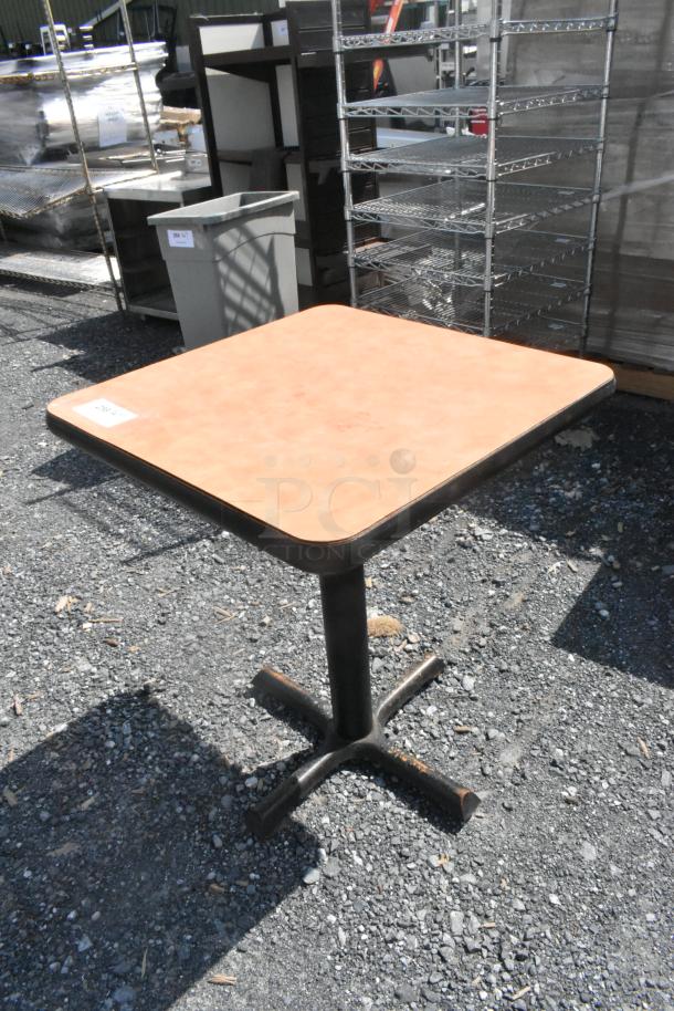 Square dining height table with light brown top and black metal base, in used condition, outdoors on gravel.