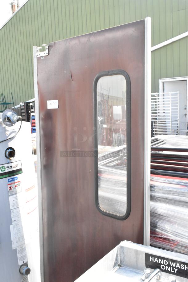 Brown swinging kitchen door with oval window, metal frame, and visible wear. Tag shows "267 N.M.". Suitable for restaurant use.