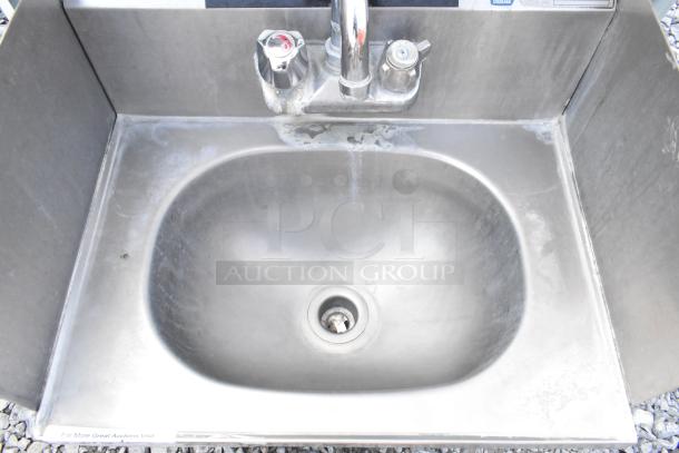 Stainless steel wall mount sink with side splash guards, faucet, and handles, showing signs of wear.