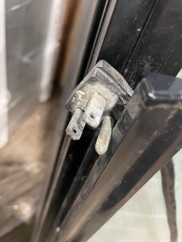 Close-up of a plug for a True GDM-23 commercial refrigerator with black frame and glass door. Visible wear on plug prongs.