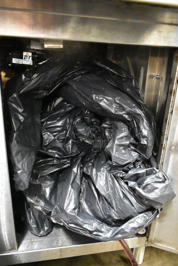 Stainless steel counter with one open door showing black garbage bags inside, under shelf visible; good condition.