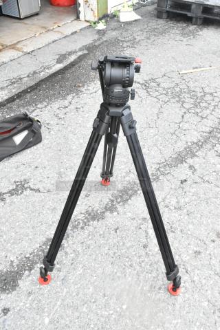 Sachtler tripod with fluid head on pavement, shown with a soft case nearby. Appears in good condition, ideal for professional video use.
