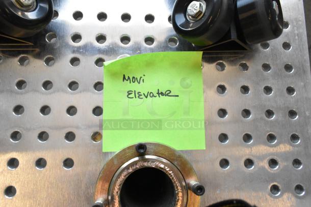 Movi Elevator on perforated metal base with black wheels, labeled with a green sticky note for identification.
