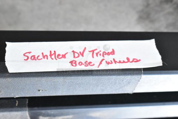 Tape with handwritten label reading "Sachtler DV Tripod Base/Wheels" on a metal surface.