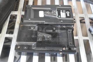 V Mount to Gold Mount Adapter, black with visible mounting screws, used condition, placed on metal grate surface.