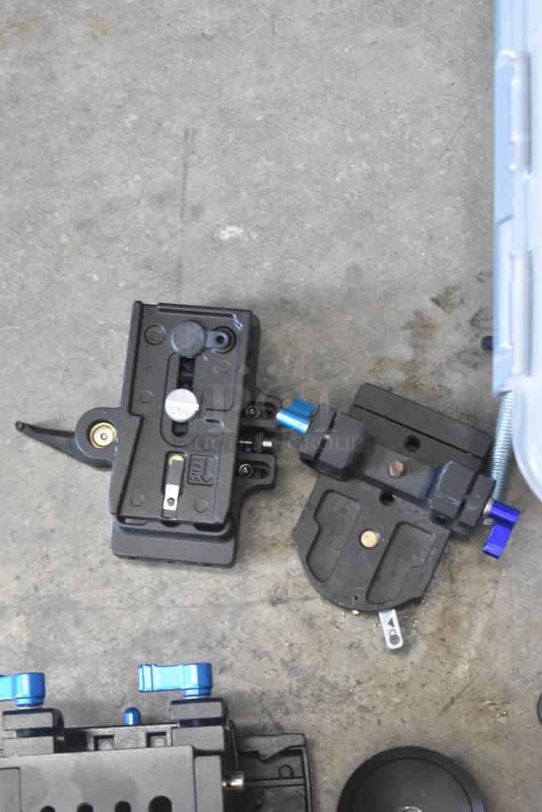 Various camera attachment gear in a poly bin, including quick-release plates and adjustment knobs, shown on concrete.
