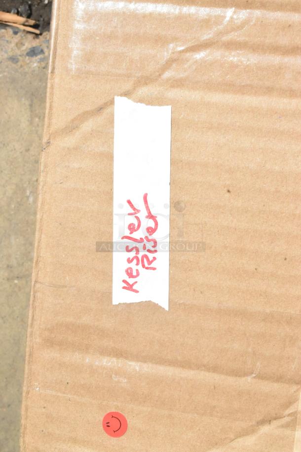 Cardboard box labeled "Kessler Riser" in red text, with a small red sticker featuring a smiley face. Box appears intact.