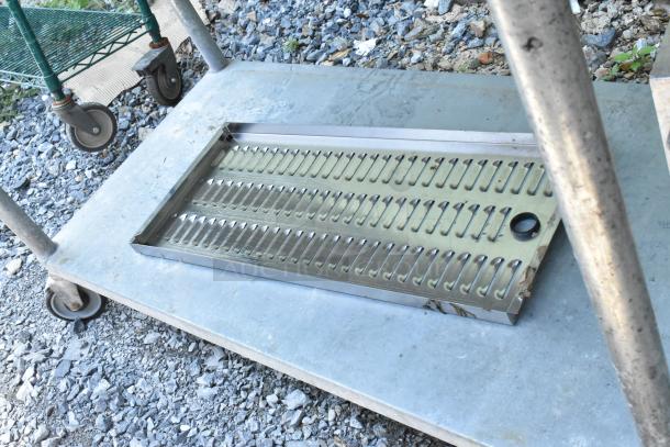 Stainless steel table with under shelf, containing dunnage racks and metal pans, showing signs of wear and outdoor use.