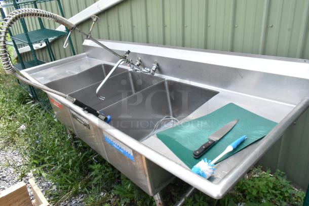 Stainless steel 3 bay sink with dual drain boards, handles, and spray nozzle attachment in used condition.