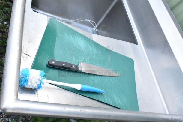 Stainless steel 3 bay sink with dual drain boards, handles, and spray nozzle attachment. Includes visible cutting board and utensils.