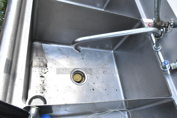 Stainless steel 3 bay sink with dual drain boards, spray nozzle, visible dirt, and cup. Equipped with hot and cold handles.