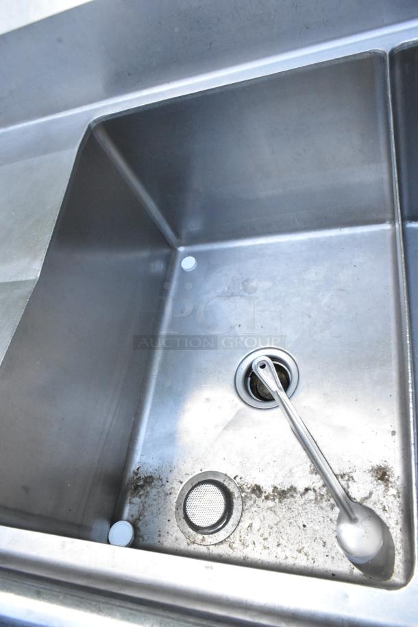 Stainless steel 3 bay sink with dual drain boards, integrated handles, and spray nozzle attachment. Signs of wear visible.