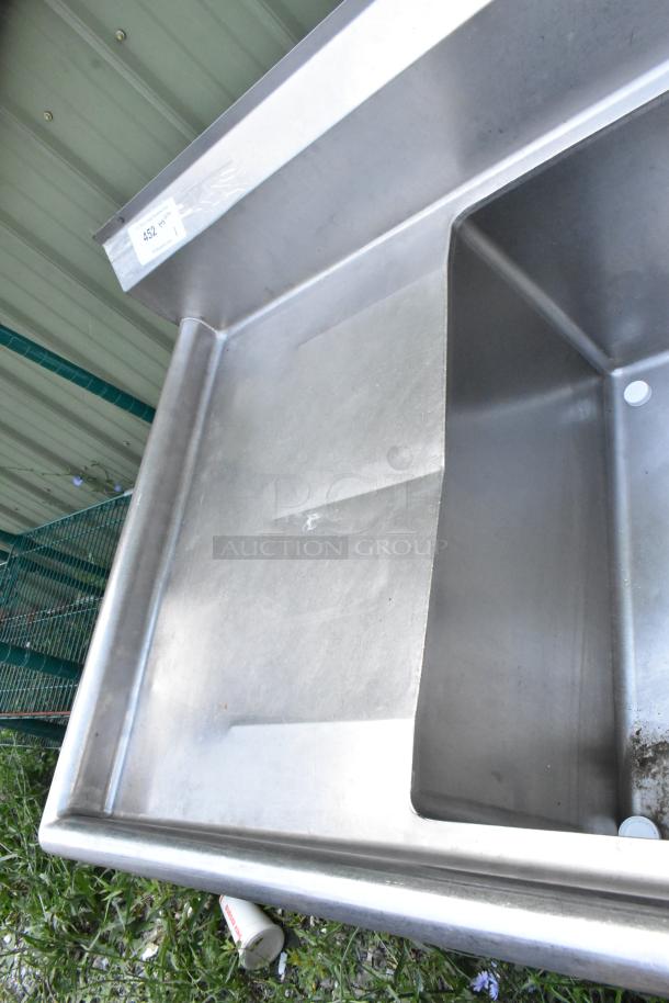 Stainless steel 3-bay sink with dual drain boards, handles, and spray nozzle; clean and polished, item tag visible on upper edge.