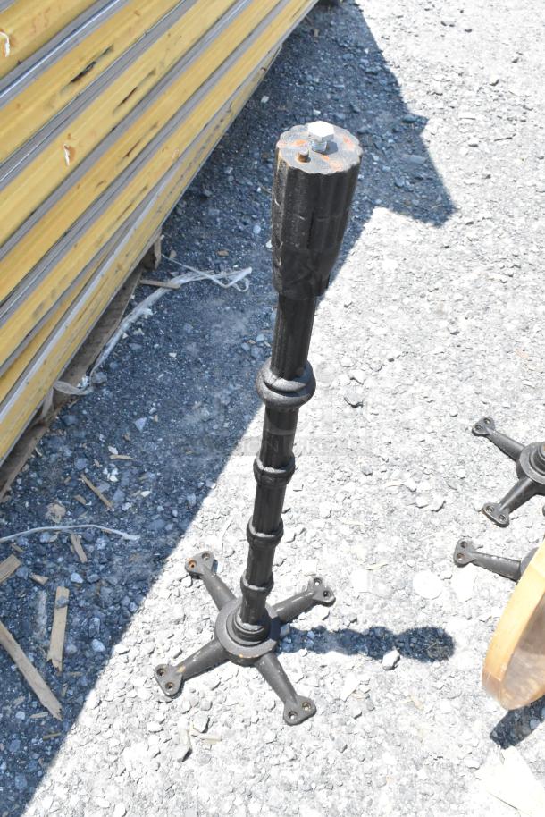 Metal table base with a cross-shaped bottom, showing signs of weathering. Sturdy construction, suitable for outdoor use.