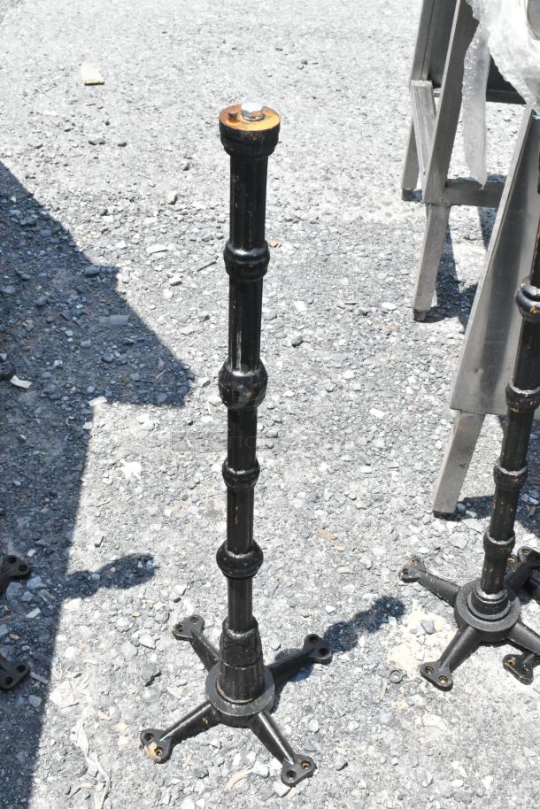 7 black metal table bases with four-footed design on gravel surface. Includes 1 tabletop. Some rust visible.