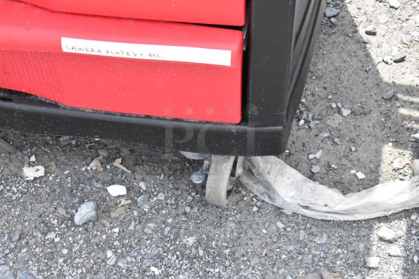 Black and red poly cart with label "Camera Plates + Acc." on commercial casters, shown on gravel surface.