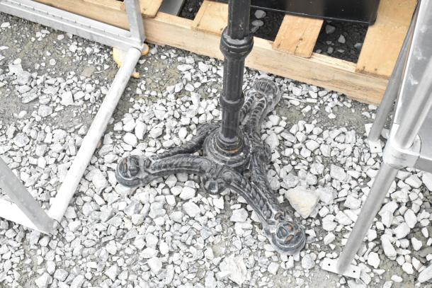 Black metal table base with ornate design, shown outdoors on gravel. Suitable for supporting various tabletop sizes.