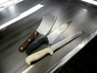 Three chef knives in great condition on stainless steel surface: a cleaver with wooden handle, a chef's knife with black handle, and a serrated knife with white handle.