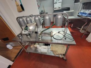 5' stainless steel work table on casters with Nemco 5-bulb warming lamp, kitchen supplies underneath. Appears functional.