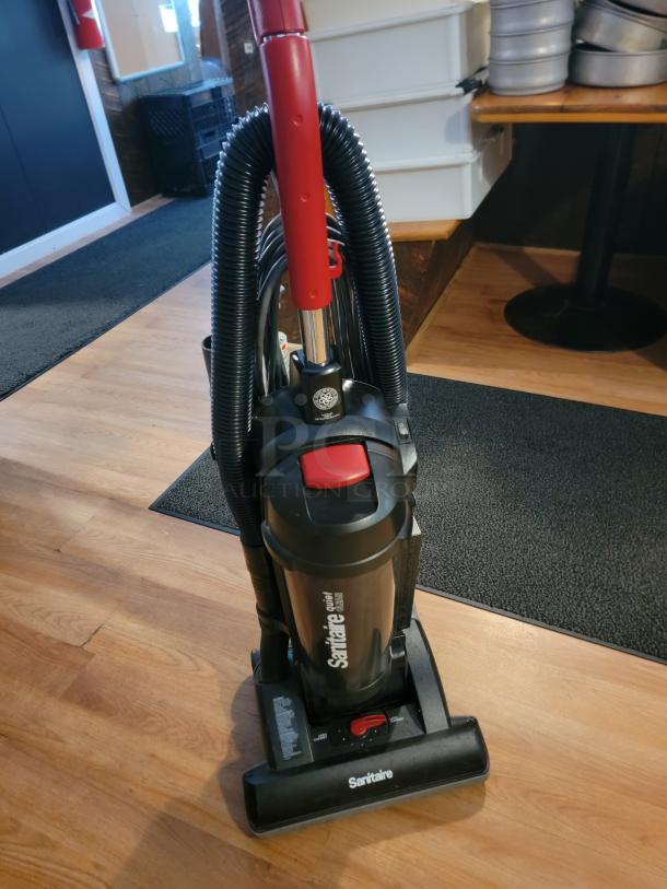 Sanitaire Quiet Clean commercial upright vacuum, displayed on a wood floor, features a red handle and black hose.