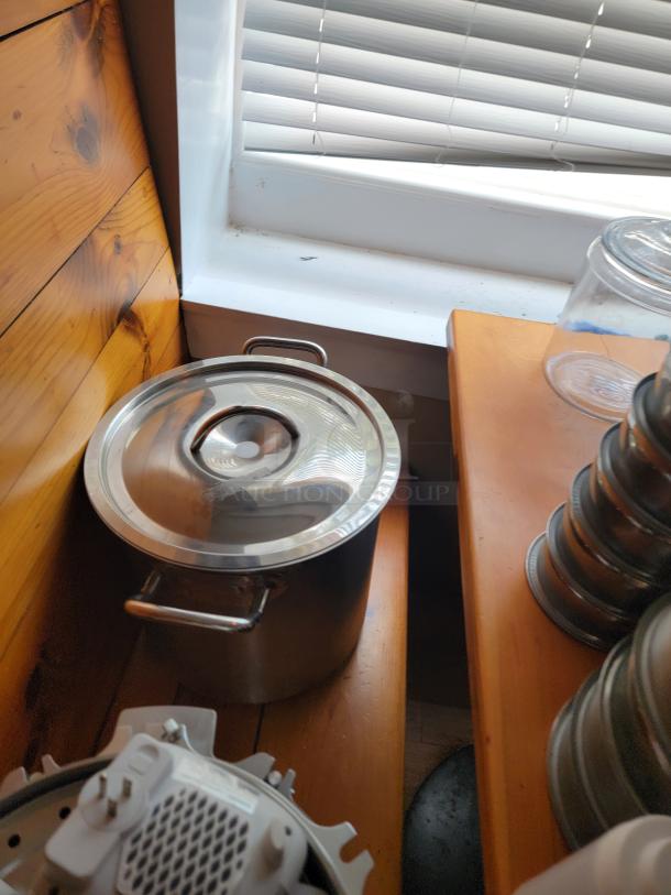 Assorted commercial kitchen supplies including a stainless steel pot with lid and stacked metal containers, shown on a wooden surface.