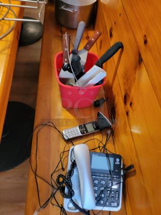 Lot of commercial kitchen supplies includes utensils in a red container, pot, and AT&T phones with cords on a wood counter.