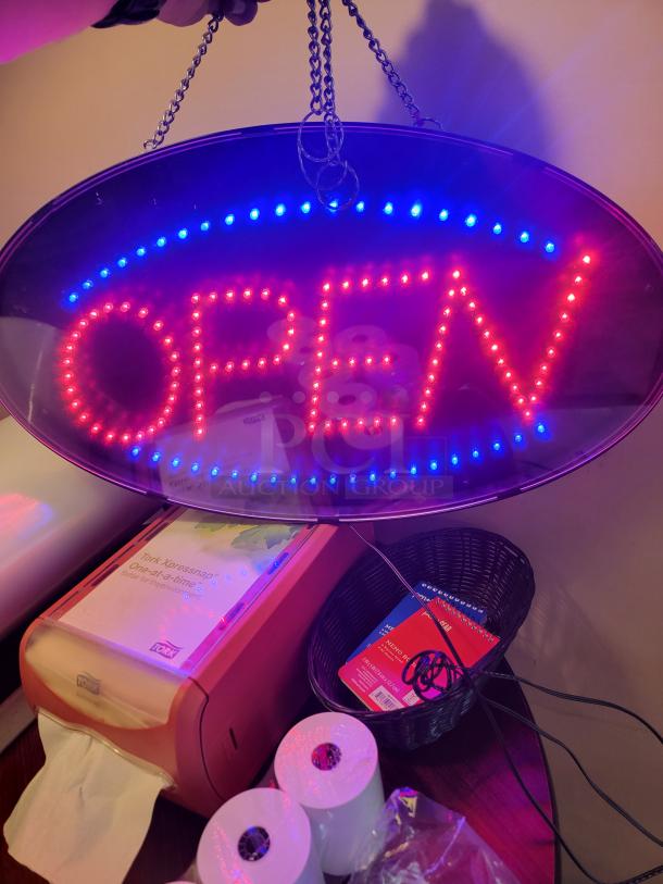 LED "OPEN" sign, oval-shaped, brightly lit with red and blue lights. Includes Tork Xpressnap napkin dispenser and extra napkin rolls. Ideal for small businesses.