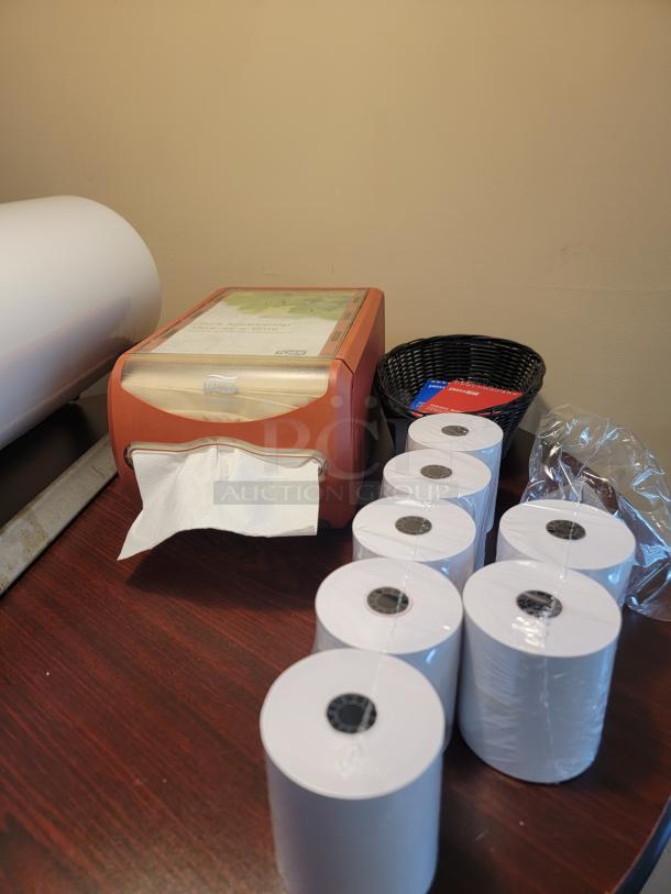 Office supplies including Tork Xpressnap dispenser, thermal paper rolls, and basket with miscellaneous items on a wooden surface.