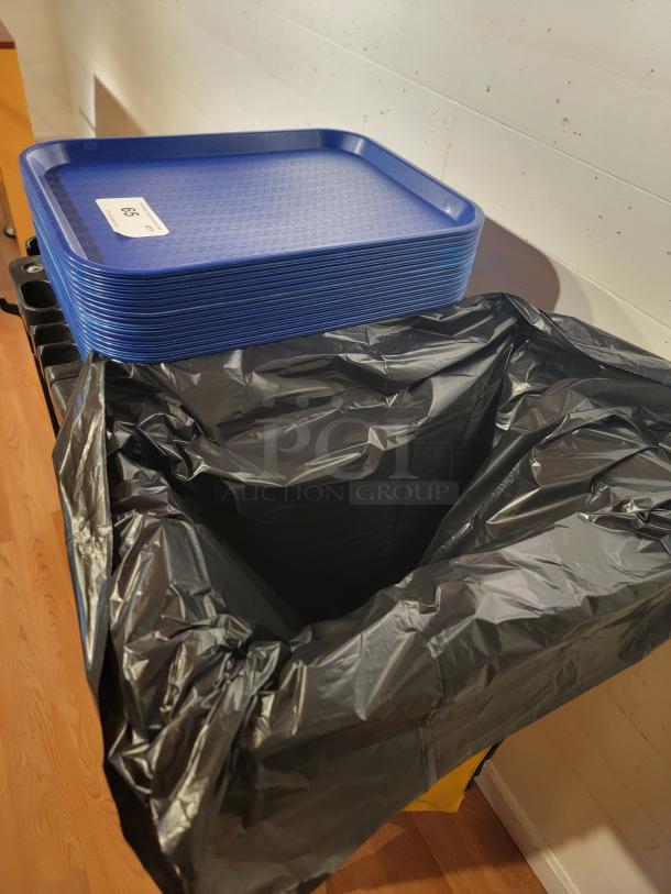 RubberMaid custodial rolling cart with trash bag holder, supply shelves, hooks, and stack of blue cafeteria trays.