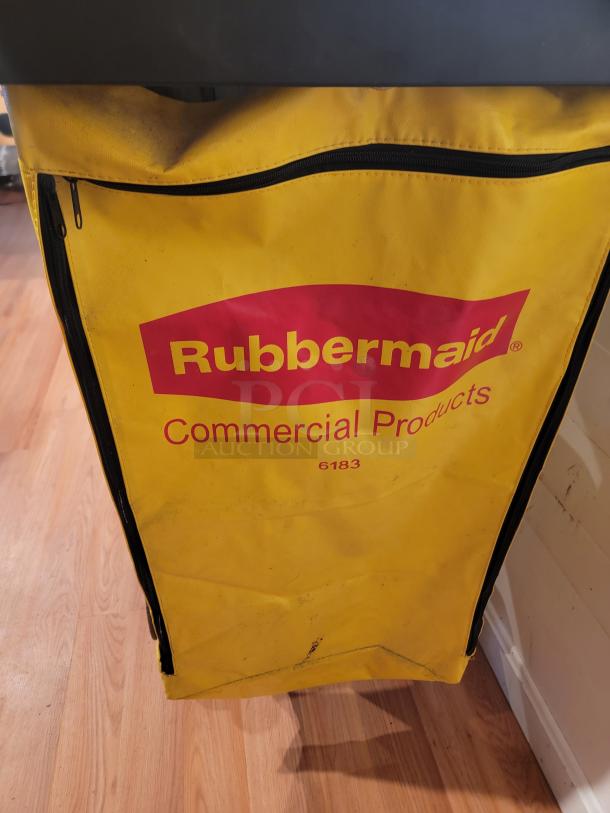 RubberMaid custodial rolling cart with attached yellow trash bag holder, featuring supply shelves, hooks, and cafeteria trays.