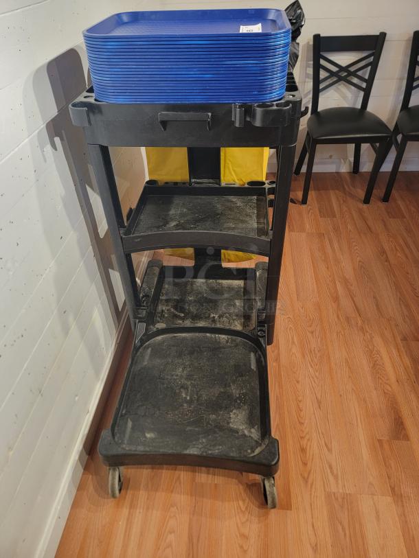 RubberMaid custodial rolling cart with shelves, trash bag holder, hooks, and stacked blue cafeteria trays. Used condition.