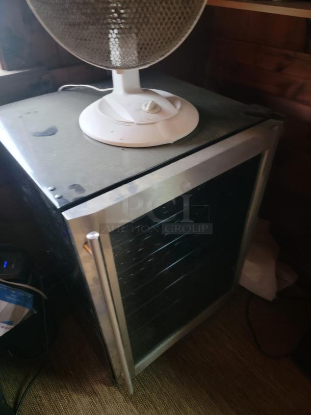 Small black mini fridge with glass front and silver handle, topped by a white oscillating fan. Good condition, ready for removal.