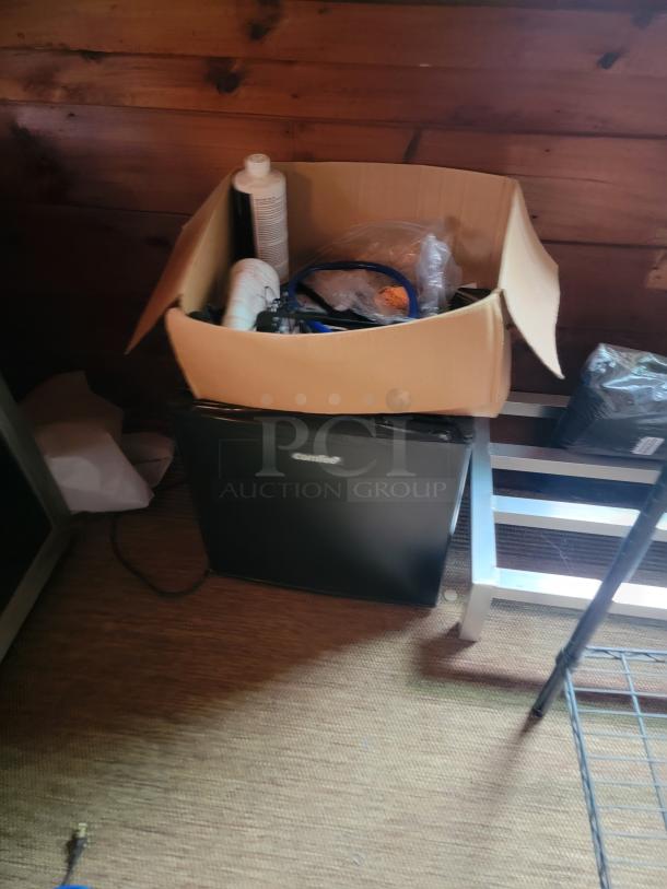 Cardboard box filled with assorted office supplies atop a black compact refrigerator. Silver metal shelf partially visible.