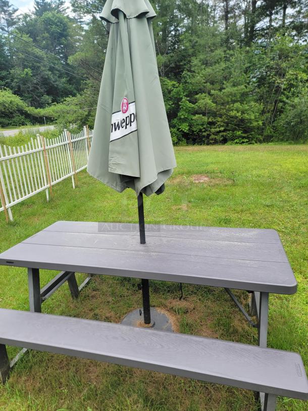 Folding picnic table with a green umbrella and weighted base on grass. Perfect for outdoor events. Umbrella shows "Schweppes" logo.