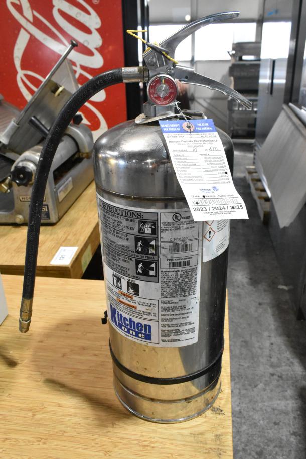Brand new Kitchen One wet chemical fire extinguisher, scratch and dent condition, stainless steel, instructional labels and inspection tag visible.