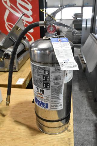 Brand new Kitchen One wet chemical fire extinguisher, scratch and dent condition, stainless steel, instructional labels and inspection tag visible.