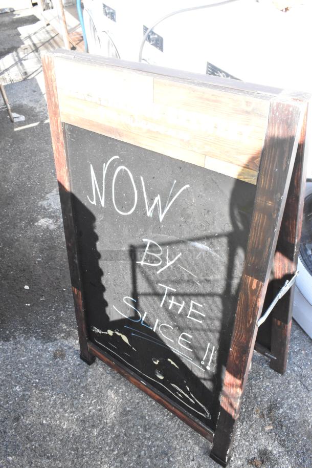 Wood pattern A-frame sidewalk sign with chalkboard surface, weathered condition. White text reads "NOW BY THE SLICE!!".