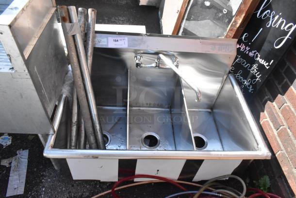 Stainless steel 3 bay sink with faucet and handles. Visible pipes and signs of wear. Auction tag marked "164". Ideal for commercial use.