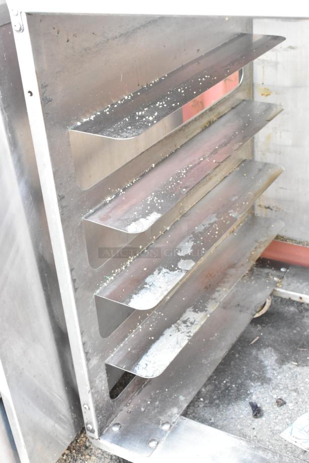 Stainless steel pan transport rack with commercial casters, showing multiple shelves. Visible wear with food debris present.