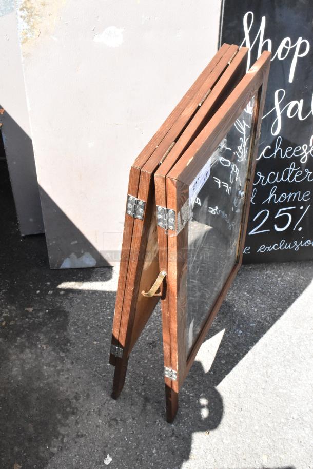 Wooden A-frame sidewalk signs, metal hinges, clear panels, slight wear. Text partially visible, suitable for businesses.