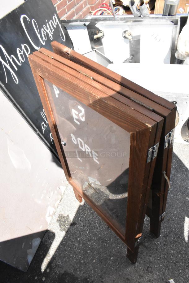 Two wooden A-frame sidewalk signs with clear panels, showing partial text. Signs have metal hinges and appear in used condition.