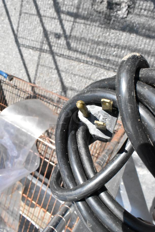 Close-up of a black coiled cable with a three-prong plug, placed atop a wire shelving unit, partially visible in the background.