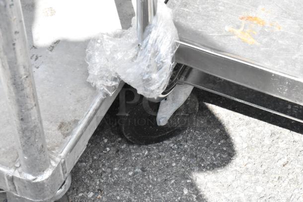 Stainless steel 2-tier cart with commercial casters; some wear, wrapped in plastic. Durable for heavy-duty use.